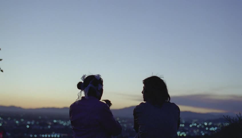 Silhouette of two people sitting and talking and handing something back and forth as the city lights shine below with mountains in the distance