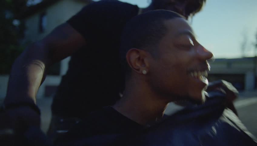 A man is sitting outside in a chair smiling and laughing as a woman puts a cape on him for a haircut
