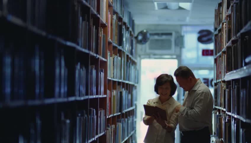 Man and woman talk and smile while sharing book between row of book shelves
