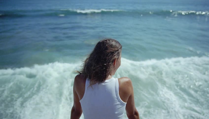 A woman is sitting on rocks along the coast as waves splash high near her face
