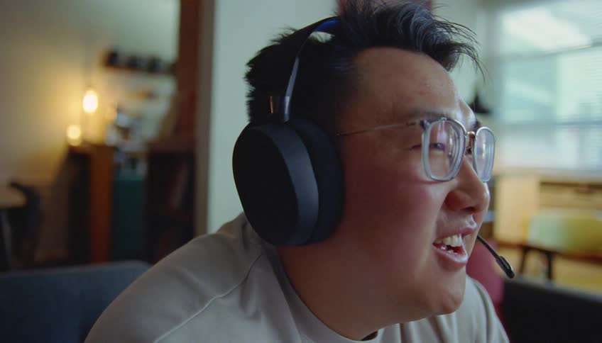 A man wearing a headset and microphone smiles and shouts while sitting playing a game