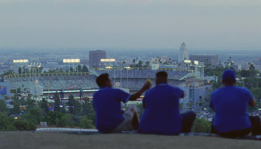 Three friends clink beer bottles as they watch a sporting event from a vantage point over the stadium
