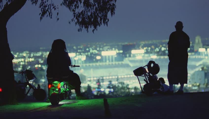 People with motorbikes standing on hill overlooking city are silhouetted against bright city lights at night