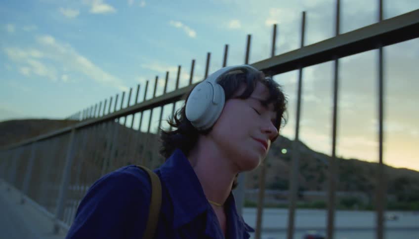 Woman with wavy hair purple shirt crossing highway overpass listening to music through headphones