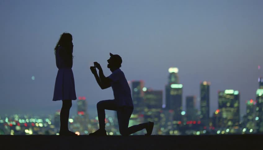 Silhouette of a man kneeling down proposing to his girlfriend in front of the city skyline at evening