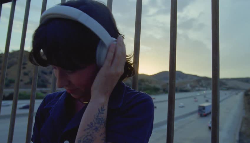Woman with wavy hair tattoo purple shirt and white headphones leans against iron fences listening to music on overpass