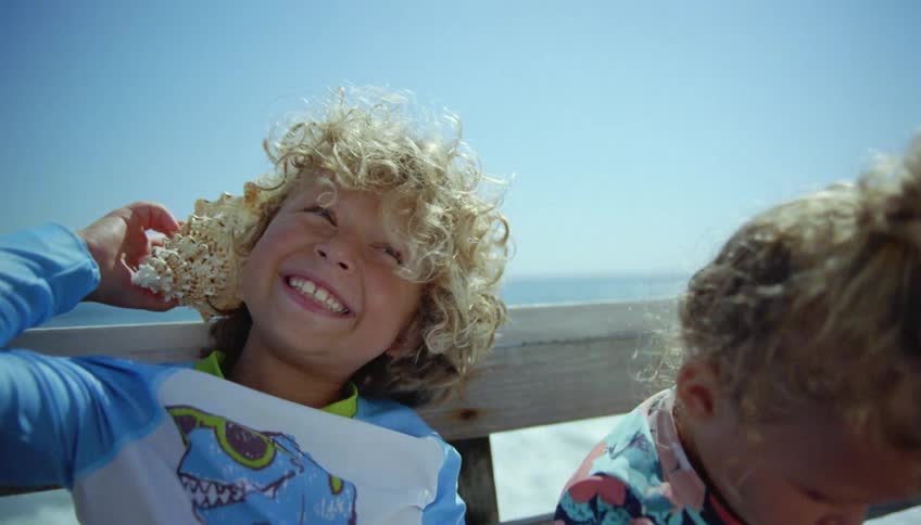 Boy with curly hair sitting next to girl listens to conch shell on the coast