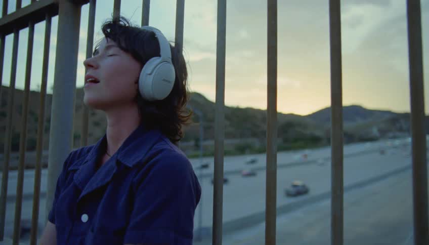 Woman listening to music on headphones leans against rail on bridge as traffic moves along highway