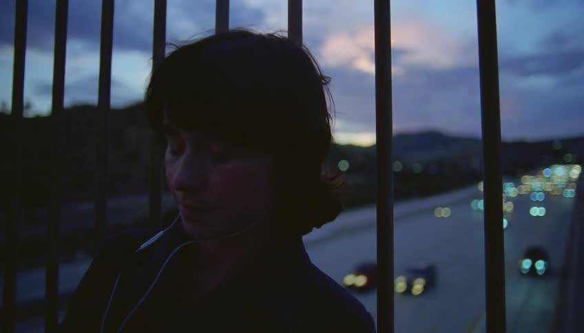 A teenage girl is sitting on a bridge listening to music in her headphones as traffic moves on the street below