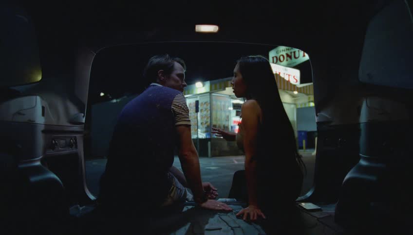 Young couple comfortably sitting on the back of a car talking in front of a donut shop at night