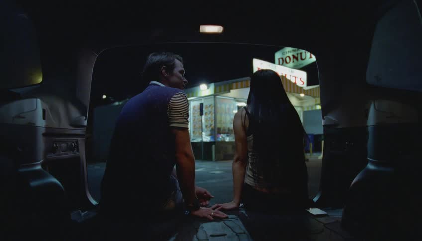 Young couple comfortably sitting on the back of a car talking in front of a donut shop at night