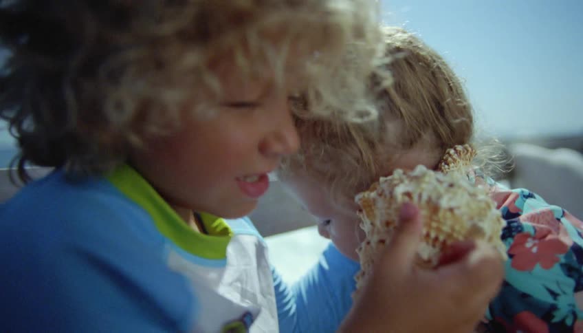 Blonde brother and sister hold spiral conch shells to their ears sitting on bench next to ocean