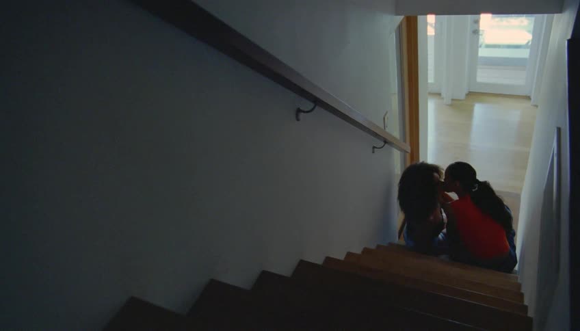 Girl with curly hair and girl with ponytail and red top kiss on the lips sitting on stairs at home