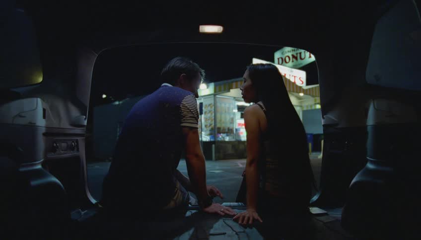 A man and a woman are sitting on a the side of a street across from a donut shop talking