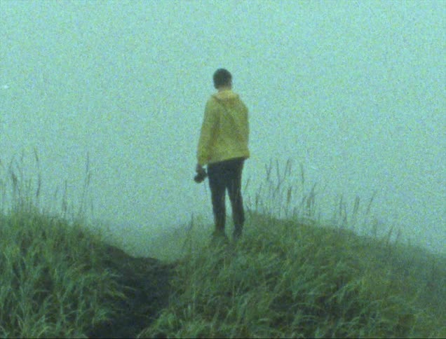 Man wearing black beanie yellow jacket and black pants holds camera standing on grass hill slope on foggy day