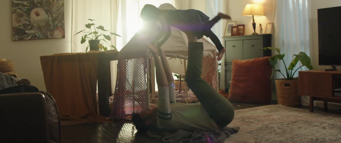 A man at home sits on the floor and holds his daughters hands and leans back and puts his feet on her stomach and lifts her off the ground and then lowers her back down