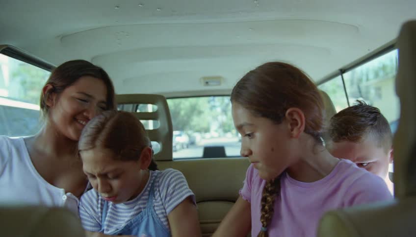 A boy sits in the back seat of a car next to his three sisters that are fighting and plays on a cell phone