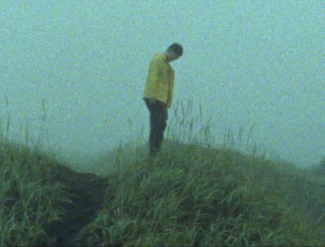 Young man wearing a bright yellow jacket stands on a small hill caught on vintage film