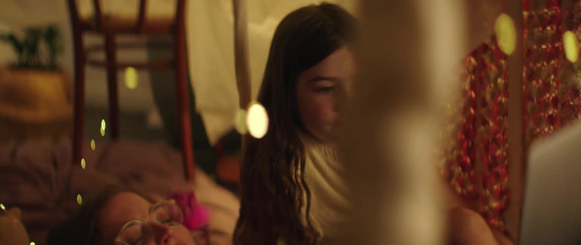 Girl with long brown hair watches laptop next to woman lying on bed in bedroom
