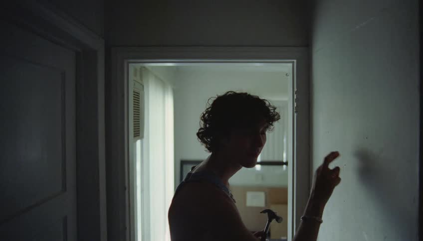 A man holding a hammer taps a hallway wall with his fingers listening for a stud