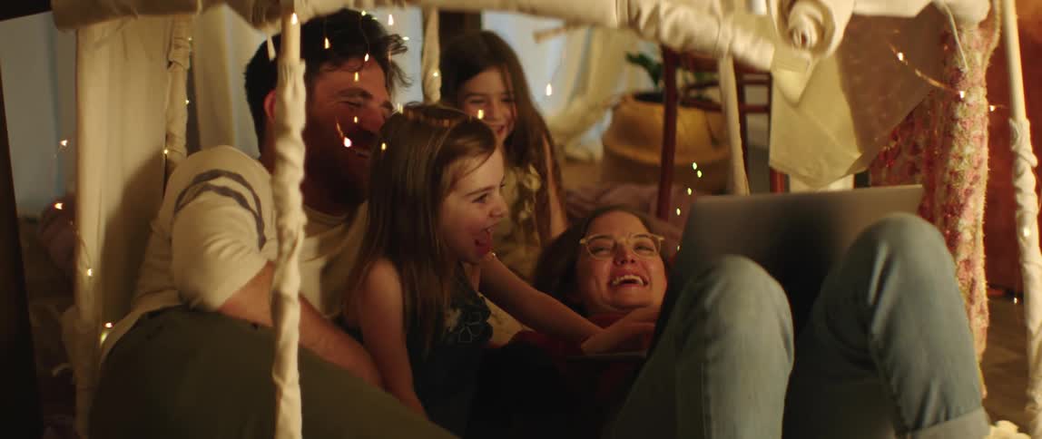A mother and father lie on the floor with their two daughters and laugh as they look at a laptop