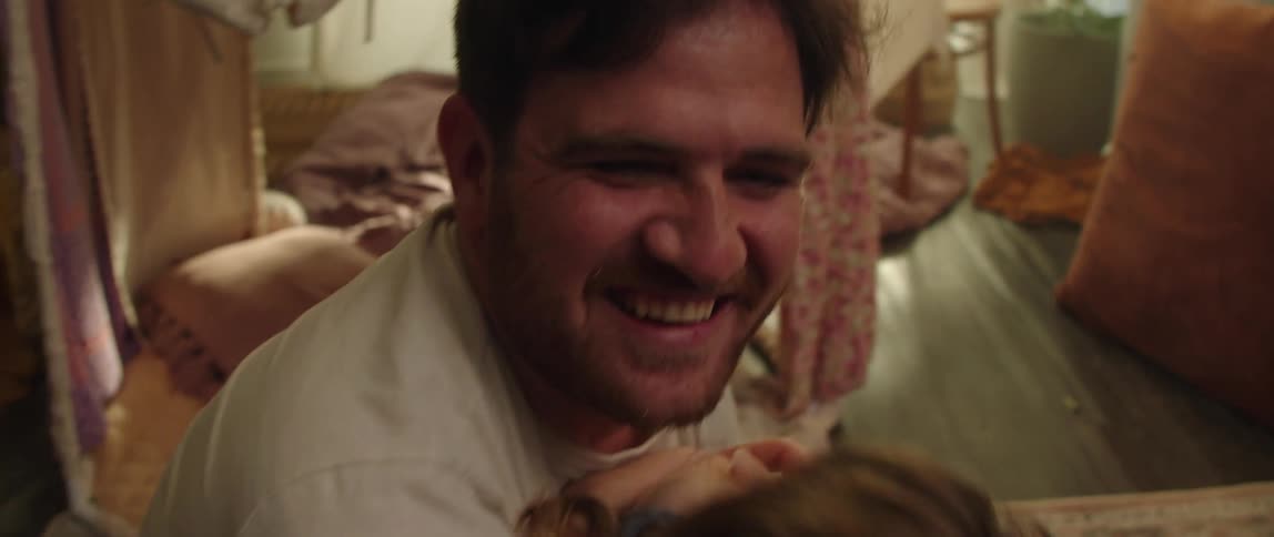 Father with beard white sweatshirt and green pants hugs daughter with brown hair and blue dress in the living room