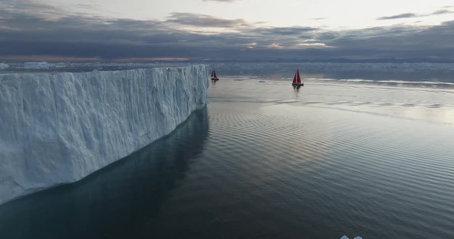 Small sailboats pass large icebergs in the ocean