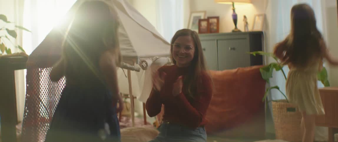 Mother with brown hair glasses and red sweatshirt plays with two girls jumping up and down in living room