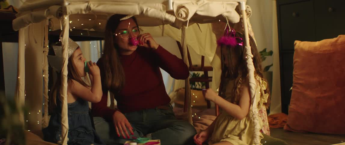 A woman sits in a fort at home and has a tea party with her two girls