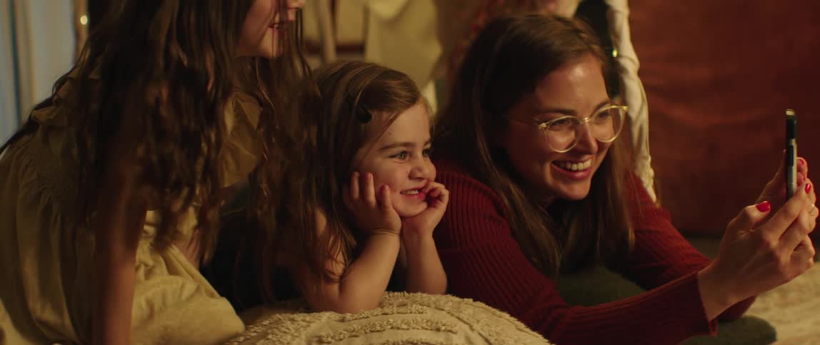 A mother lies with her two daughters and they laugh as she shows them her cell phone as she scrolls on it
