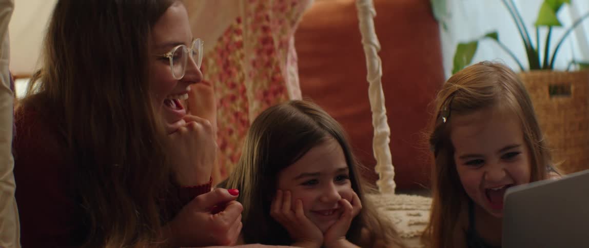 Mother and daughters laugh while lying on bed and watching laptop