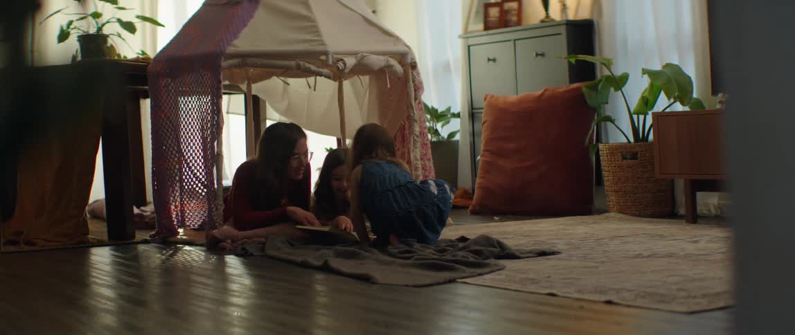 Mother laying in the floor of the living room with her daughters reading stories on a book