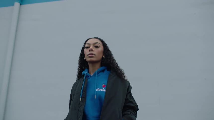 A young woman is wearing a jacket as she stands outside of a building with neighborhood houses across the street