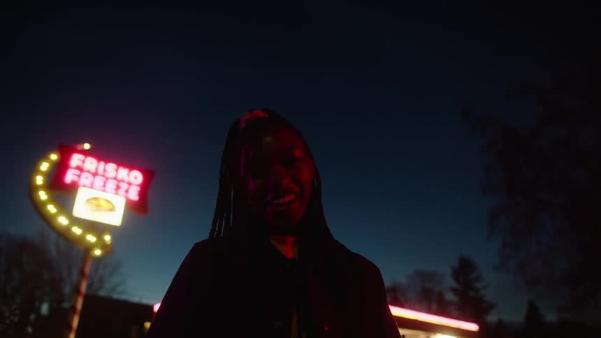 A teenage girl stands laughing and smiling in the frisko freeze parking lot where a group of friends has gathered