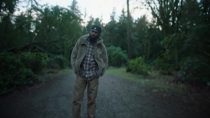 A man stands with his hands in his pockets on a gravel road in the woods