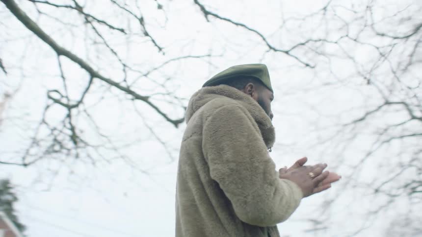 A man is standing outside near bare trees rubbing his hands together in the cold