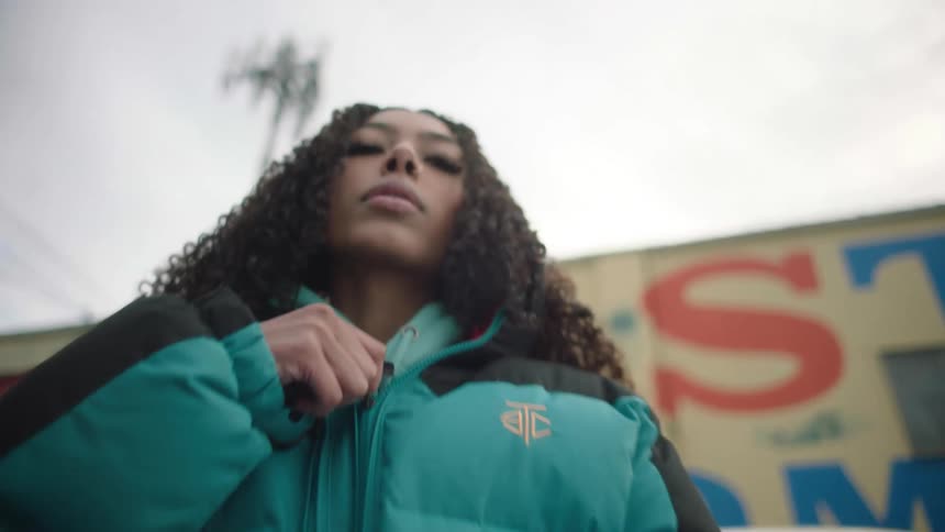Young woman reclined on a car parked in front of a small store zips up her jacket while looking at the camera