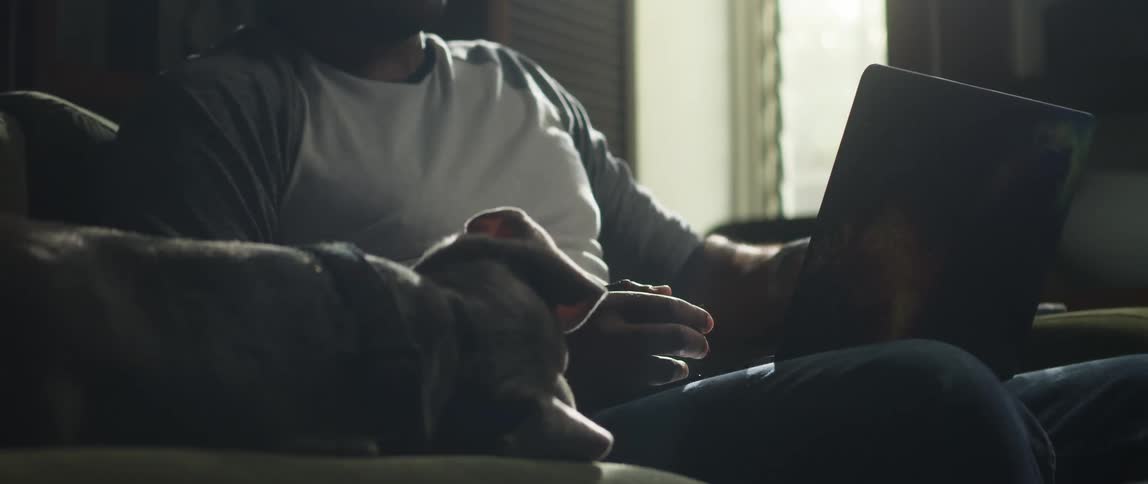 A man sitting on the couch sets down his cell phone and types on a laptop computer as his dog is lying next to him