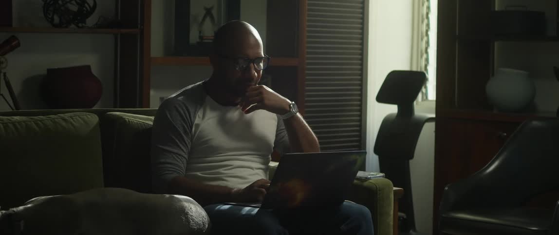 A man is sitting on his living room couch using a laptop as his dog is lying next to him