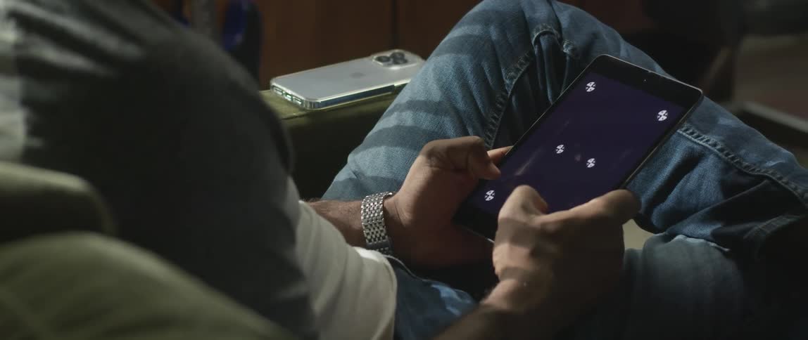 Man sits tapping on midsized mobile device while sitting near smartphone on arm of furniture