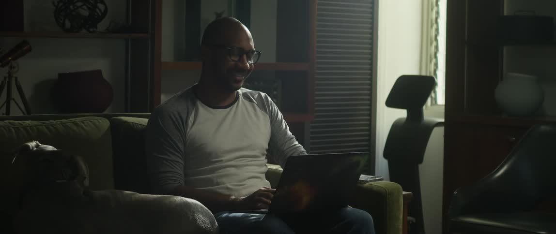 A man sits on his couch next to his dog and laughs as he looks at the laptop in his lap