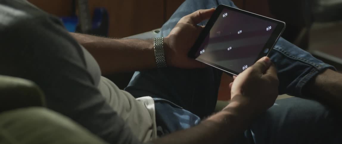 Man with sweatshirt silver wristwatch and jeans sits down on green couch holding tablet
