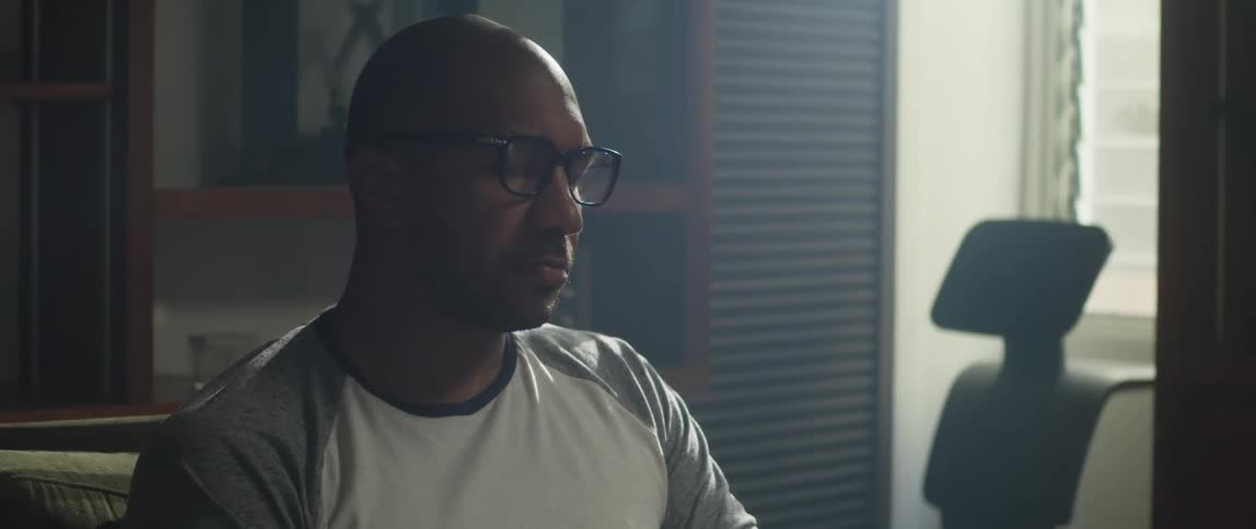 Bored man with shaved head glasses and sweatshirt sits at desk sighing