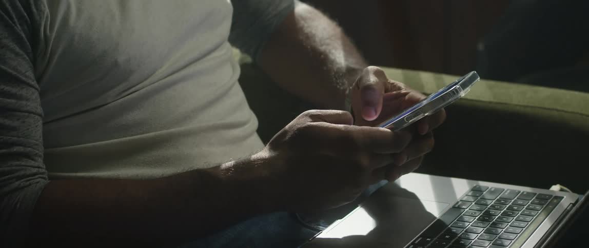 A man with a laptop in his lap types and swipes on his cell phone