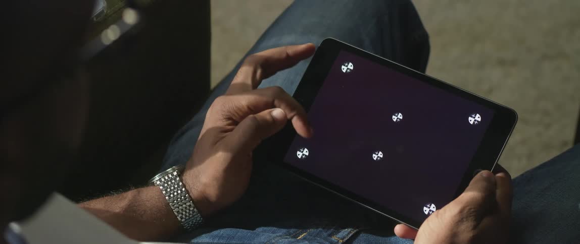 A man is sitting down holding a tablet with white symbols on the screen and he moves his finger across the screen