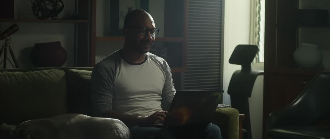 A man is sitting on his living room couch typing on a laptop computer as his dog is lying next to him