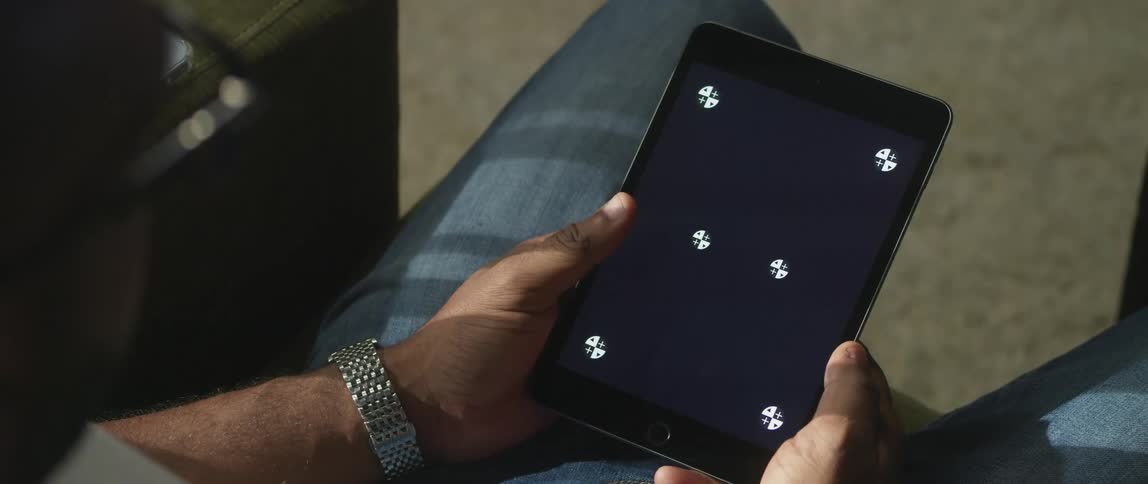 A man is sitting down holding a tablet with white symbols on the screen and he moves his thumb across the screen