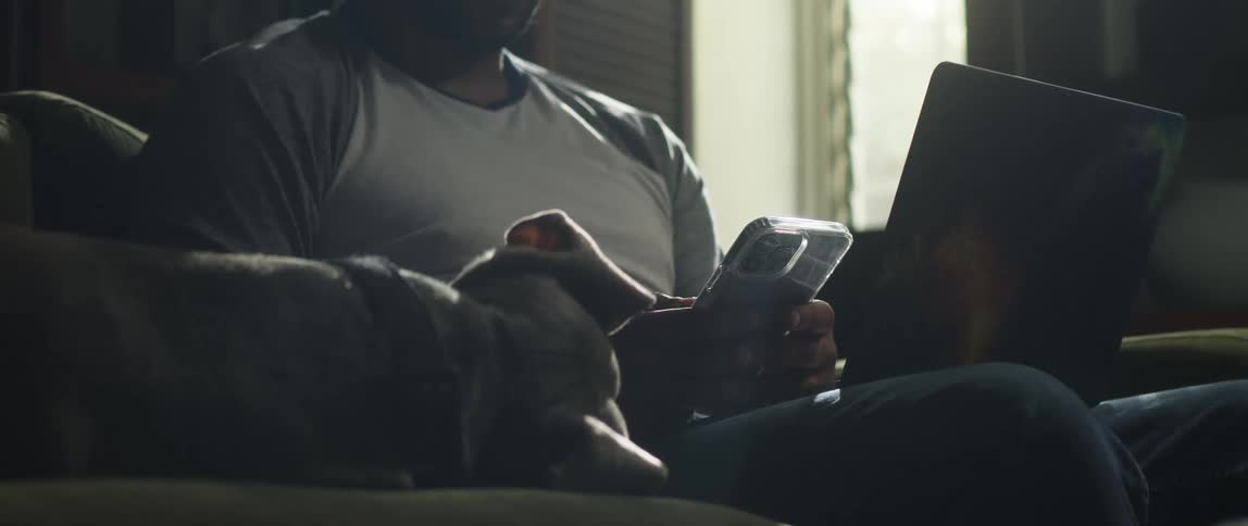 Man sits on couch using a smartphone and laptop while a dog sleeps next to him