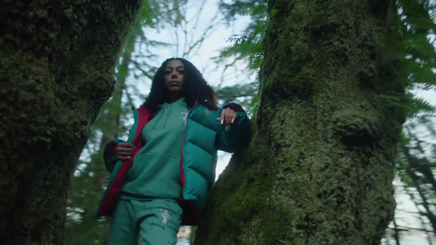 Woman with curly hair turquoise coat top and pants stands between tree trunks in the woods