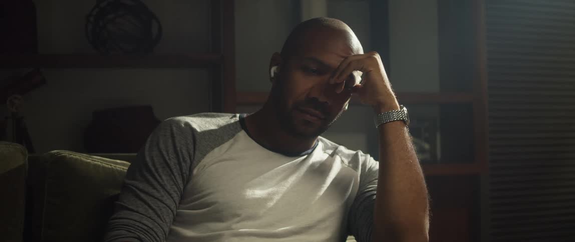 A sad looking man with earbuds in sits on a couch at home and rests his head in his hand